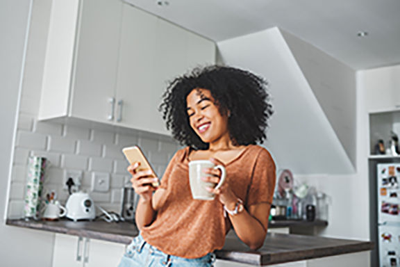 Woman in kitchen looking at phone holding coffee mug